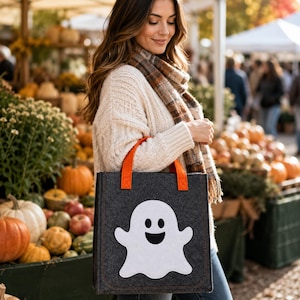 Könnte beinhalten: Eine dunkelgraue Filztasche mit einem großen weißen Geistermotiv und einem schwarzen Gesicht. Die Tasche hat zwei leuchtend orangefarbene Stoffgriffe an der Oberkante. Die rechteckige Tasche besteht aus dickem, strukturiertem Filzmaterial.