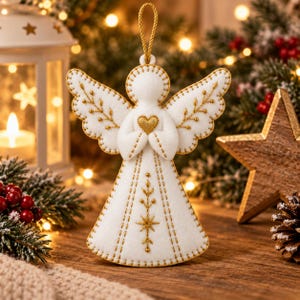 May include: A white felt angel ornament featuring gold embroidered details on the wings and dress. The angel holds a small gold heart at the center of its chest and includes a gold hanging loop at the top for display on a tree or hook.