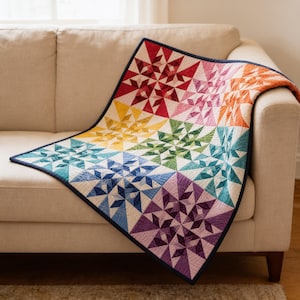May include: A quilt featuring a vibrant rainbow color gradient arranged in a star pattern. The design consists of multiple square blocks in shades of red, orange, yellow, green, blue, and purple, all finished with a dark blue binding edge.