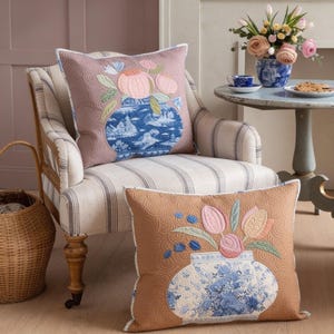 Könnte beinhalten: Zwei quadratische Zierkissen mit gesteppten Oberflächen und Stickereien von blau-weißen Vasen, gefüllt mit rosa und pfirsichfarbenen Blumen. Ein Kissen ist malvenfarben, das andere hellbraun, beide mit detaillierten, geschwungenen Steppmustern.