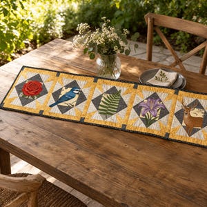 May include: A rectangular quilted table runner featuring five distinct square panels with nature-themed illustrations including a red rose, a blue bird, a green fern leaf, a purple lily, and a brown deer, all bordered by yellow and black fabric.