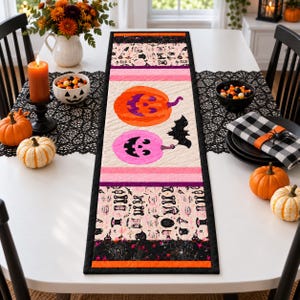 Op de afbeelding: Een rechthoekige, doorgestikte tafelloper met een crèmekleurig middenpaneel met een oranje pompoen, een roze pompoen en een zwarte vleermuis. De randen zijn voorzien van Halloween-motieven, waaronder schedels en spinnenwebben in zwart, oranje en roze.