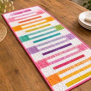 May include: A rectangular quilted table runner featuring a vibrant rainbow-colored geometric bar pattern on a white background with a pink border. The fabric displays various shades of red, orange, yellow, green, blue, and purple in a structured layout.