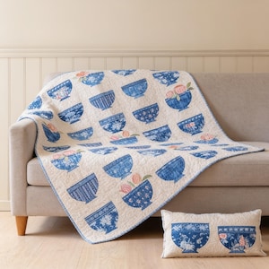 May include: A cream-colored quilt and a matching rectangular throw pillow featuring a repeating pattern of blue and white bowls. Some bowls contain illustrations of pink flowers. The quilt has a blue border and a quilted texture throughout.
