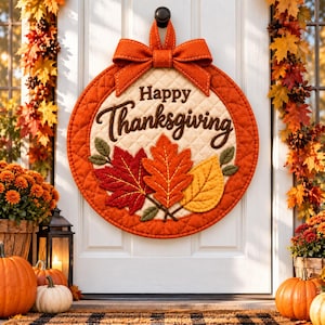 以下が含まれることがあります： Happy Thanksgivingの文字がダークブラウンの筆記体で刺繍された、円形のキルティング生地の壁掛け。中央には赤、オレンジ、黄色の刺繍された秋の葉が3枚あり、上部には吊り下げ用の大きなオレンジ色の布製リボンが付いています。