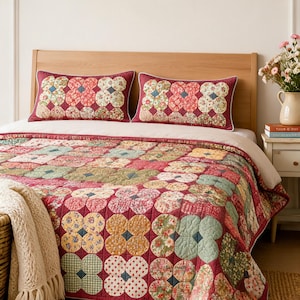 May include: A quilted bedspread and two matching rectangular pillows featuring a repeating pattern of circular floral and gingham patches in shades of red, green, yellow, and white, with a deep red background and white piping trim.