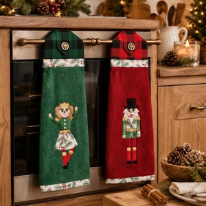 May include: Two hanging kitchen towels featuring embroidered nutcracker figures. One towel is green with a nutcracker dancer, the other is red with a soldier nutcracker. Both have plaid tops, decorative buttons, and pine branch patterned trim.