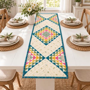May include: A rectangular quilted table runner featuring a geometric pattern of diamonds and triangles in shades of pink, yellow, green, and teal. The fabric has a cream background with small floral prints and a dark teal border trim.