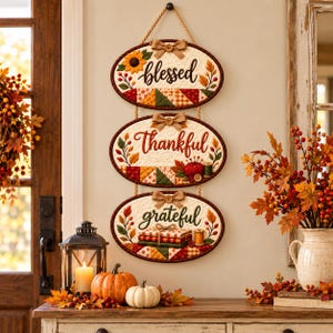 May include: A vertical wall hanging featuring three oval quilted plaques connected by rope. Each plaque has a cream-coloured centre with embroidered text reading blessed, thankful, and grateful, accented with fabric sunflowers, leaves, and buttons.