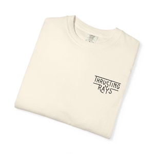 May include: A cream-colored short-sleeve t-shirt with a crew neckline. The shirt features black text on the right side of the chest that reads "THRUSTING RAYS" in a stylized, hand-drawn font. The garment has a visible interior neck label.
