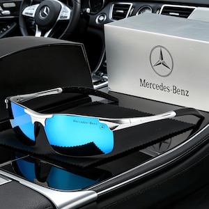 Może przedstawiać: Srebrne okulary przeciwsłoneczne z niebieskimi lustrzanymi soczewkami. Okulary mają czarną gumowaną część na zausznikach. Tekst "Mercedes-Benz" jest wydrukowany na soczewce i zauszniku. W tle srebrne pudełko z logo Mercedes-Benz.