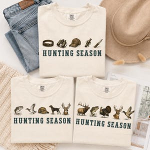 May include: Three cream-colored t-shirts featuring the text "HUNTING SEASON" in dark green. The top shirt displays a belt, binoculars, cap, flask, and knife. The bottom left shirt shows a fish, duck, cap, dog, and deer. The bottom right shirt shows an elk, fish, turkey, deer, and duck.