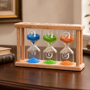 May include: A wooden frame holding three glass hourglasses with blue, green, and orange sand. Each hourglass is labeled with 3, 5, and 10 minutes respectively, featuring white circular arrows and text printed on the glass bulbs.