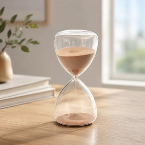 Könnte beinhalten: Eine Sanduhr aus klarem Glas mit einer abgerundeten, bauchigen Form und einem schmalen Mittelhals. Der obere Teil ist teilweise mit feinem, hellbraunem Sand gefüllt, der langsam durch die Mitte in den unteren Teil fließt und einen kleinen Haufen bildet.