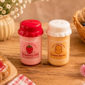 Peut inclure: Deux petits saupoudreurs en plastique avec couvercles. L'un est rose avec un couvercle rouge, étiqueté Strawberry Jelly avec un dessin de fraise. L'autre est orange clair avec un couvercle blanc, étiqueté Peanut Butter avec un dessin de cacahuète.