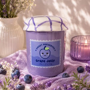 Peut inclure: Un pot en céramique violet cylindrique avec un couvercle blanc à motif quadrillé violet. L'étiquette indique Sweet Preserves, Grape Jelly, et du texte japonais avec une illustration de raisin. Le pot est entouré de myrtilles et de lavande.