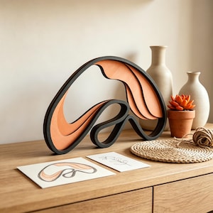 Può includere: Una scultura decorativa composta da forme in legno curve e sovrapposte, di colore nero e arancione chiaro. L'oggetto è esposto insieme a due piccoli cartoncini bianchi con disegni a linee, una piccola succulenta arancione e un tappetino intrecciato rotondo.