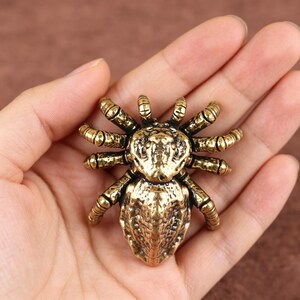 May include: A metallic gold-toned spider figurine featuring a textured, segmented body and eight curved legs. The surface of the spider displays a weathered, antique finish with dark recessed areas that highlight the intricate details of the exoskeleton.