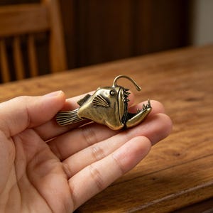 May include: A small, metallic, brass-colored anglerfish figurine with a detailed textured surface, prominent sharp teeth, a distinct dorsal fin, and a curved lure extending from its head, captured in a side profile view with a weathered finish.