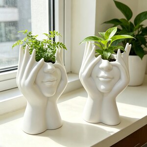 May include: Two white ceramic planters shaped like a pair of hands cupping a human face. Each planter features a smooth, matte finish and holds a small green plant. The design depicts fingers resting near the eyes and nose of the face.