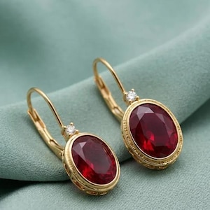 May include: A pair of gold leverback earrings featuring oval-cut deep red gemstones set in textured gold frames. Each earring is accented with a single small, clear round-cut stone positioned at the top where the gemstone meets the gold hook.