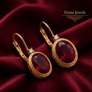 May include: A pair of gold leverback earrings featuring oval-cut deep red gemstones surrounded by a textured gold border and topped with a small clear round stone. The text Fiona Jewels is visible in the upper right corner of the image.