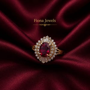 May include: A gold ring featuring a large oval-cut deep red gemstone at the center, surrounded by a halo of small round diamonds and accented with baguette-cut diamonds at the top and bottom. The text Fiona Jewels is visible above the ring.