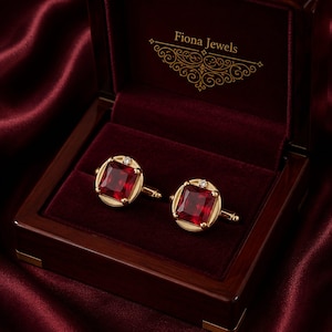 May include: A pair of gold cufflinks featuring square-cut red gemstones and small accent diamonds. The cufflinks are presented in a dark wood box with a deep red velvet interior, featuring the gold-printed text Fiona Jewels on the lid.