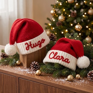 Könnte beinhalten: Zwei rot-weiße Strick-Weihnachtsmützen mit geripptem Umschlag. Die größere Mütze ist mit dem Namen Hugo in roter Schreibschrift bestickt, die kleinere mit dem Namen Clara in roter Schreibschrift. Jede Mütze hat einen weißen Bommel an der Spitze.