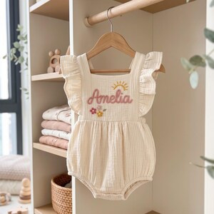 Könnte beinhalten: Ein cremefarbener Musselin-Strampler mit Rüschenärmeln und quadratischem Ausschnitt. Die Vorderseite ist mit dem Namen Amelia in Rosa, einer gelben Sonne und kleinen bunten Blumen bestickt. Das Kleidungsstück hängt an einem Holzkleiderbügel.
