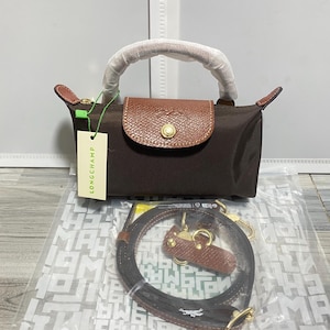 May include: A brown handbag with a brown leather flap and handle. The bag has a gold-colored clasp and a detachable shoulder strap. A tag with the word "LONGCHAMP" is attached to the bag.