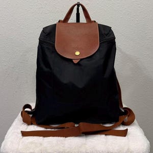 May include: Black backpack with a brown leather flap and handle. The flap is secured with a gold-colored button. The backpack has brown shoulder straps and a brown strap at the bottom. The bag is sitting on a white surface.
