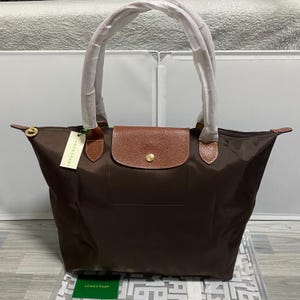 May include: A brown tote bag with a brown leather flap and gold-tone hardware. The bag has long, white handles and a small tag that reads "LONGCHAMP". The bag is made of a durable material and is suitable for everyday use.