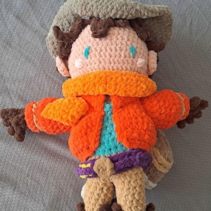 May include: A handmade crochet doll featuring a tan face, brown hair, and a wide-brimmed tan hat. The figure wears a bright orange jacket over a turquoise shirt, a purple belt with a tan buckle, tan pants, and dark brown boots with a tan bag.