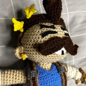 May include: A crocheted doll featuring a tan face, a prominent dark brown mustache, and dark brown hair with a tuft on top. The figure wears blue overalls with brown straps and has yellow star-shaped accents near the hair and a small yellow earring.