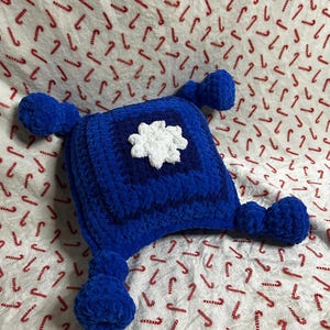 May include: A square, blue, crocheted plush pillow featuring a textured, dark blue center square and a white, six-petaled flower applique in the middle. The pillow has four rounded, blue, crocheted appendages extending from each corner.