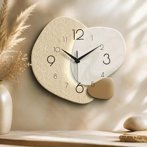 Könnte beinhalten: Eine abstrakte Wanduhr mit einer großen, strukturierten beigen Basis in Halbmondform, überlagert von einem glatten, weiß marmorierten Abschnitt und einem kleinen, einfarbigen hellbraunen Akzentstück. Sie hat schwarze Zeiger und Ziffern von 1 bis 12.