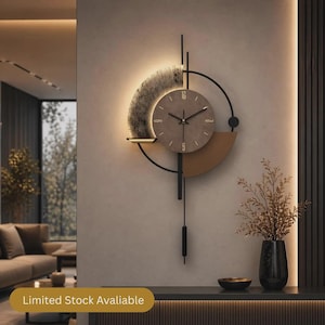 Op de afbeelding: Een wandklok met een ronde bruine wijzerplaat met gouden cijfers op 12, 3, 6 en 9 uur. De klok is omlijst door een zwarte metalen structuur met een textuurrijk halfrond accent, geïntegreerde achtergrondverlichting en een verticaal pendelonderdeel.