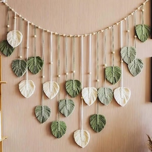 Puede incluir: Un colgante de pared de macramé con diseños en forma de hojas en tonos verdes y crema. Las hojas están suspendidas de una cuerda con cuentas, creando una exhibición decorativa y texturizada. Esta pieza artesanal añade un toque natural y bohemio.