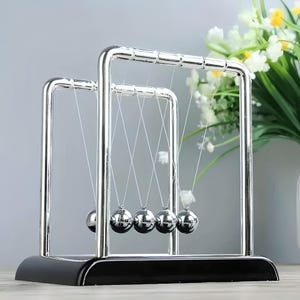 May include: A Newton's cradle desk toy with five polished steel balls suspended from a chrome frame. The base is black, and the balls hang in a row, demonstrating the transfer of momentum. The background includes a vase of white flowers.