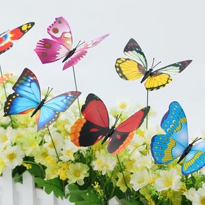 Può includere: Una collezione di decorazioni da giardino a forma di farfalla colorata. Le farfalle presentano vari motivi alari in tonalità di rosso, rosa, giallo e blu. Sono attaccate a sottili paletti neri, adatte per la decorazione esterna tra i fiori.