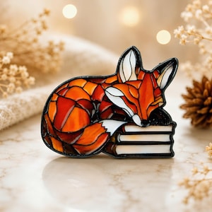 Pode incluir: Um broche decorativo com um design de raposa em estilo vitral em tons de laranja, vermelho e branco, apoiando a cabeça numa pilha de três livros. A raposa tem contornos pretos a separar os segmentos coloridos e a ponta da cauda branca.