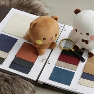 May include: A fabric swatch sample book featuring various textured squares in shades of cream, navy, charcoal, rust, and tan. A brown plush bear and a white plush bear with brown ears sit on the pages next to a handheld magnifying glass.