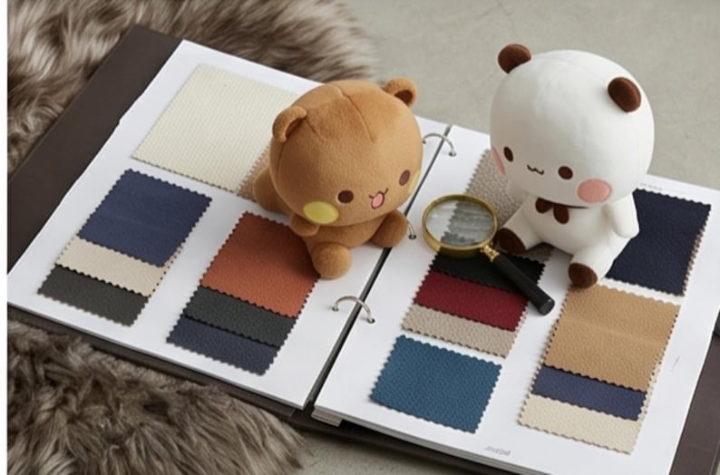 May include: A fabric swatch sample book featuring various textured squares in shades of cream, navy, charcoal, rust, and tan. A brown plush bear and a white plush bear with brown ears sit on the pages next to a handheld magnifying glass.