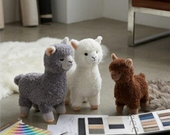 May include: Three plush alpaca toys in grey, white, and brown with textured, curly fur. The toys stand upright with small ears and dark eyes. Nearby are a color swatch fan, a pair of scissors, a sketch on paper, and a fabric sample book.