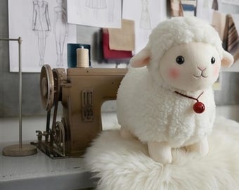 May include: A plush white lamb toy with a textured, curly fleece body, smooth face, pink cheeks, and black eyes. The lamb wears a thin brown cord necklace featuring a small, round, shiny red bell. The toy is positioned next to a sewing machine.