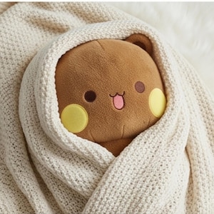 May include: A brown plush toy with a rounded head, small ears, and a face featuring dark brown eyes, yellow circular cheeks, and a pink mouth with a tongue sticking out, wrapped snugly in a textured, cream-colored knitted fabric blanket.