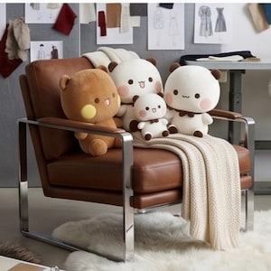 May include: A brown leather armchair with a chrome frame features four plush bear-like stuffed toys in white and brown colors. A cream-colored knitted throw blanket is draped over the seat, partially covering the cushion and the armrest.
