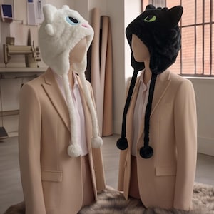 May include: Two plush animal-themed hats with ear flaps and dangling pom-poms. One hat is white with large blue eyes and a pink nose, while the other is black with vibrant green eyes. Both hats are displayed on mannequins wearing beige blazers.