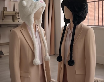 May include: Two plush animal-themed hats with ear flaps and dangling pom-poms. One hat is white with large blue eyes and a pink nose, while the other is black with vibrant green eyes. Both hats are displayed on mannequins wearing beige blazers.